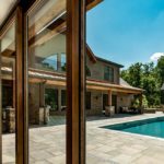 Maison moderne avec grande baie vitrée, terrasse, piscine et pavillon en bois, entourée d’arbres.